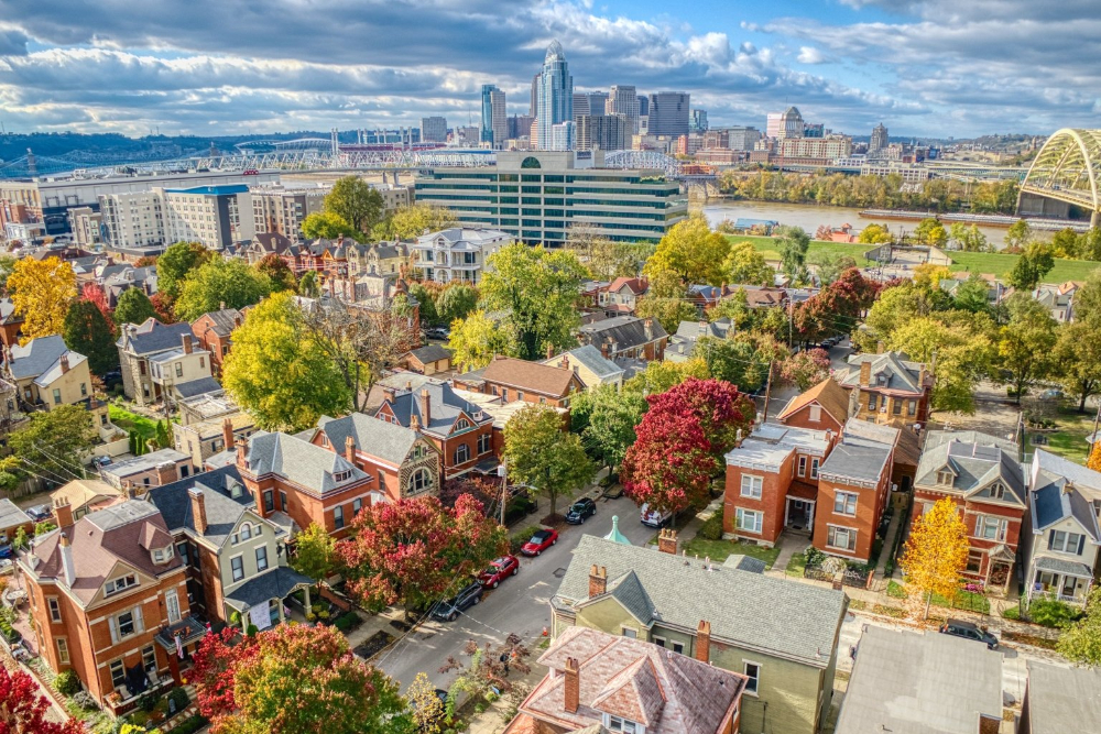 Aerial view of a autumn-colored residential neighborhood with brick houses, trees, and a city skyline across a river.