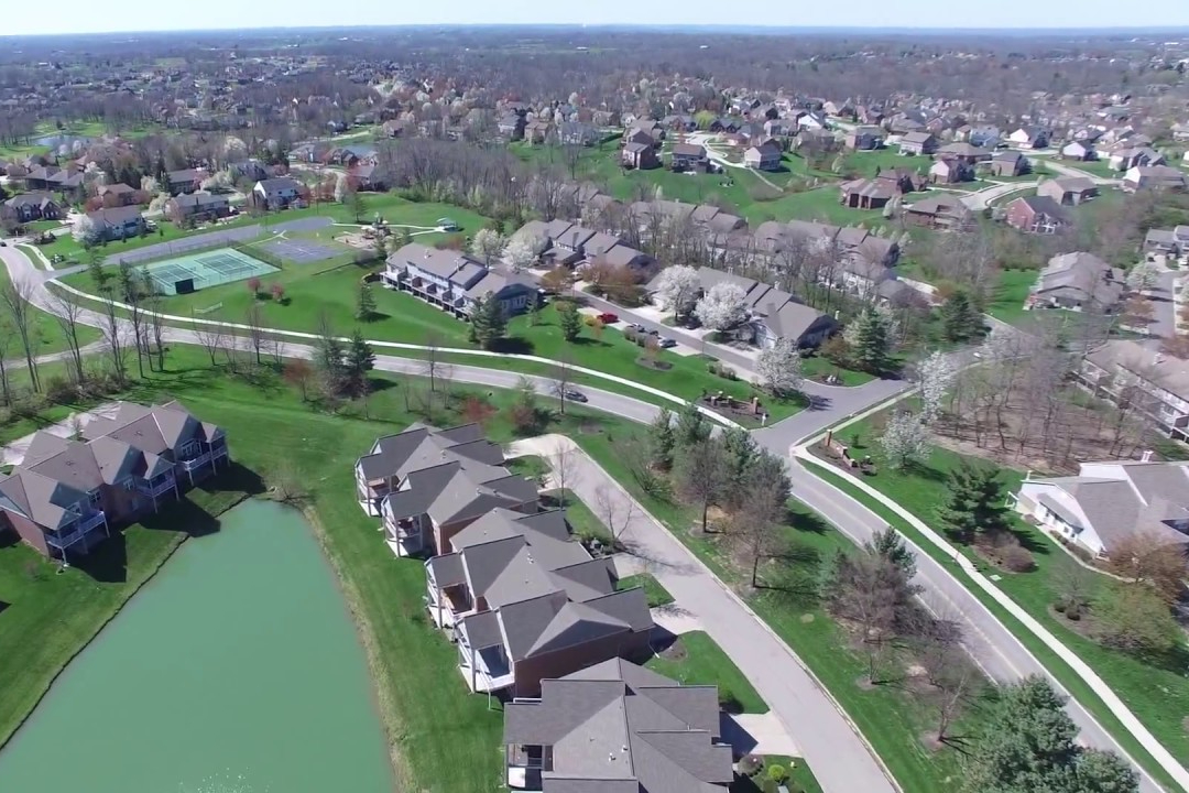 Aerial view of a suburban housing development with a curved road, row of townhomes near a pond, and green lawns.",