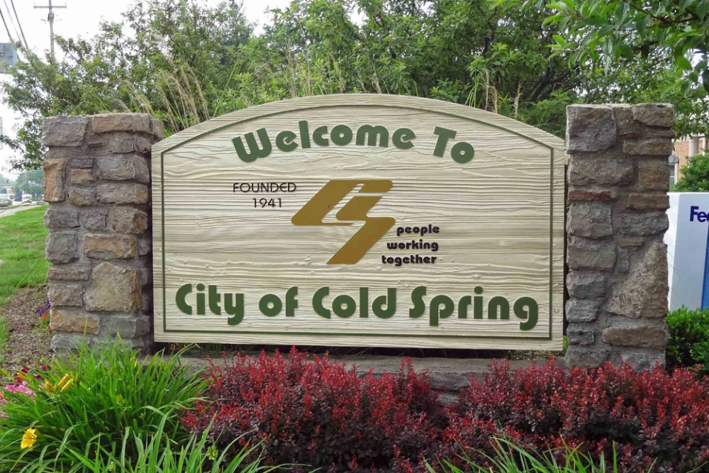 Wooden welcome sign flanked by stone pillars reading 'Welcome To City of Cold Spring' with greenery around it.