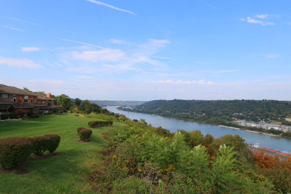 Overlooking a river valley with a manicured lawn and townhouses on the left, hills across the water under a blue sky.