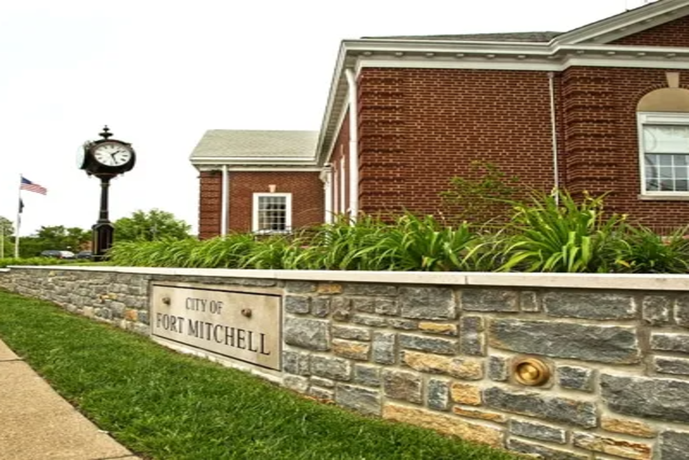 Ceremonial stone wall with a engraved sign reading 'City of Fort Mitchell' in front of a brick building and clock tower nearby, American flag in the background