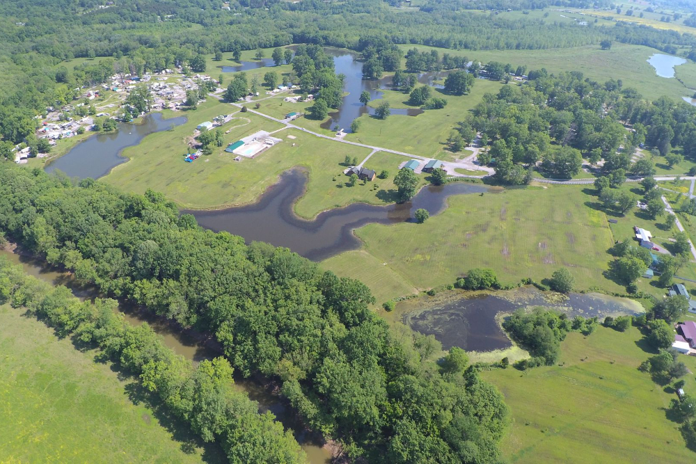 Aerial view of a park with winding ponds, grassy fields, and trees, plus scattered buildings and a campground area in the distance.