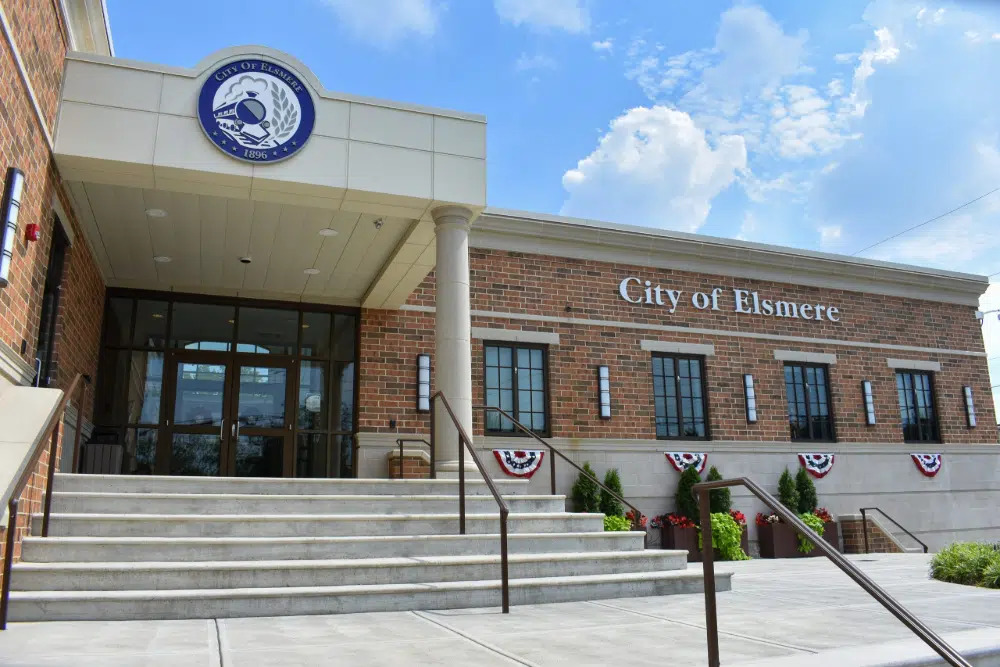 Front view of the City of Elsmere building: brick facade, stairs to double doors, and a seal above the entrance with patriotic bunting along the railing.