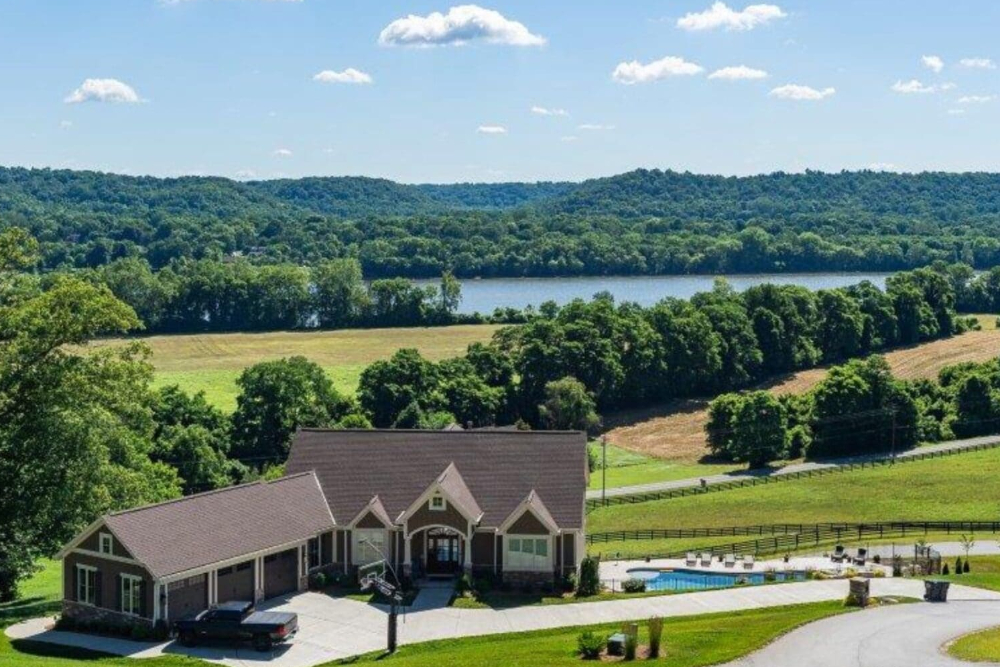 A ranch-style house with a brown roof sits on a grassy slope next to a circular driveway and a swimming pool in front, with rolling fields, trees, and a lake in the distance on a sunny day.