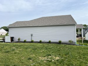 Side of a pale siding house with a steep roof, green lawn, and garden hose on the ground in the foreground.