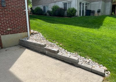 Concrete block seating wall with a pebble bed along the patio edge, a green lawn behind it, and houses in the background in a residential yard.