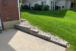Patio with a low stacked-block wall and pebble edge along a grassy slope, brick house on the left and a beige building with sliding doors in the background.