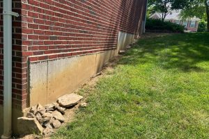 Side view of a brick building with a concrete foundation along a grassy slope; loose rocks piled at the base of the wall, and a downspout on the left.