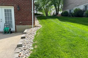 Brick house side with white door, green garden hose on a wheeled reel, and a rock border along a concrete path by a lush lawn.