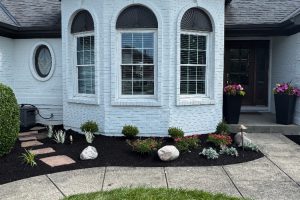 Front of a light blue brick house with a bay window, mulched flower bed, stepping stones, and potted flowers near the entryway.