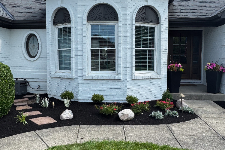 Light blue brick house with a three-panel bay window, a dark front door, and two tall planters with pink flowers at the entry; landscaped front yard with mulch, rocks, and stepping stones.