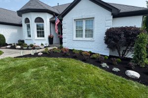 Front yard of a white brick house with an American flag near the entrance, a mulched garden bed with colorful flowers and shrubs, decorative rocks, and a curved concrete walkway. A green lawn is in the foreground.