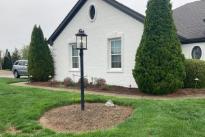 White brick house with two windows and a circular vent, a black lantern-style post in a mulched garden bed, and tall evergreen trees nearby. A silver SUV is parked in the driveway.