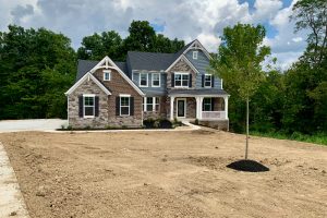 Front view of a newly built two-story house with stone and siding, a small porch, and a dirt yard awaiting landscaping with a young tree in the foreground.