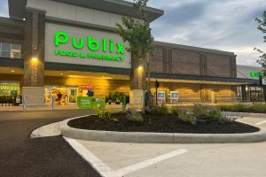 Publix storefront with bright green 'Publix Food & Pharmacy' sign, entering customers, and a landscaped curb in front.