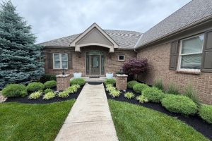 Brick house front with arched porch, dark entry door, and manicured shrubs along a concrete walkway to the door.