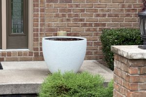 Large round light-gray planter on a concrete front porch in front of a brick wall. Soil visible inside the pot, with greenery and shrubs nearby.
